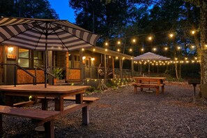 A rustic outdoor seating area with wooden benches and tables, surrounded by a building with a wooden facade and string lights. - Cedar Creek Crossing (all 4 units) (Mena)