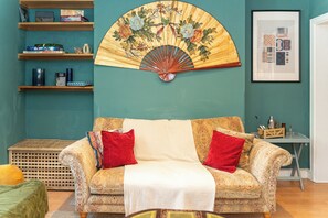 Un salon avec un canapé à motifs, un ventilateur et des étagères remplies de divers objets.