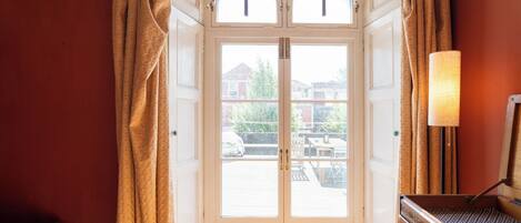 A room with a white door, two windows, and a lamp on a wooden side table.