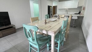 Ein Essbereich mit Holztisch und türkisfarbenen Stühlen, eine Küche im Hintergrund und ein Fernseher links.