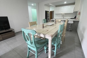 A dining area with a wooden table and turquoise chairs, a kitchen in the background, and a television on the left. - Departamento Minerva (Guadalajara)