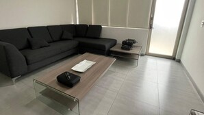Ein modernes Wohnzimmer mit einem Ecksofa, einem Glastisch, einem kleinen Beistelltisch mit einer Schreibmaschine und einer Schiebetür zum Balkon.