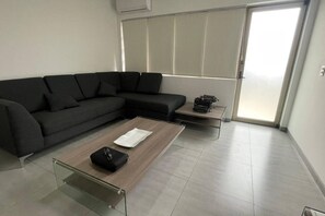 A modern living room with a sectional sofa, a glass coffee table, a small side table with a typewriter, and a sliding glass door leading to a balcony. - Departamento Minerva (Guadalajara)