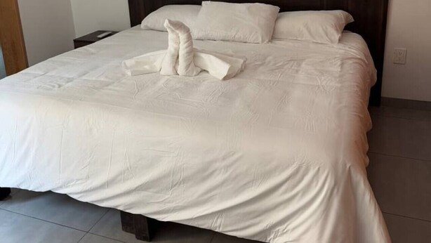 Ein ordentlich bezogenes Bett mit dunklem Kopfteil, weißen Bettwäsche und einem auf dem Bett drapierten Handtuch.