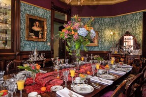Comedor formal con una larga mesa dispuesta para una comida, rodeado de muebles ornamentados y un gran centro de mesa floral.