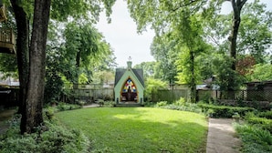 A small, green-roofed structure with a pointed arch and a stained glass window, surrounded by a well-maintained garden and a wooden fence.
