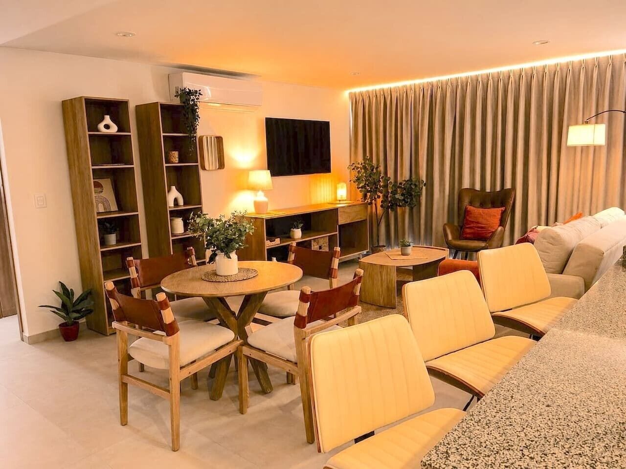 A modern living room with a round dining table, wooden chairs, a sofa, and a TV mounted on the wall.