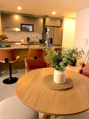 Een ronde houten eettafel met een geweven placemat, een witte vaas met groene bladeren en een moderne keuken op de achtergrond.