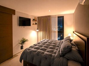 Een slaapkamer met een bed, een aan de muur bevestigde televisie, een potplant en een raam met gordijnen.