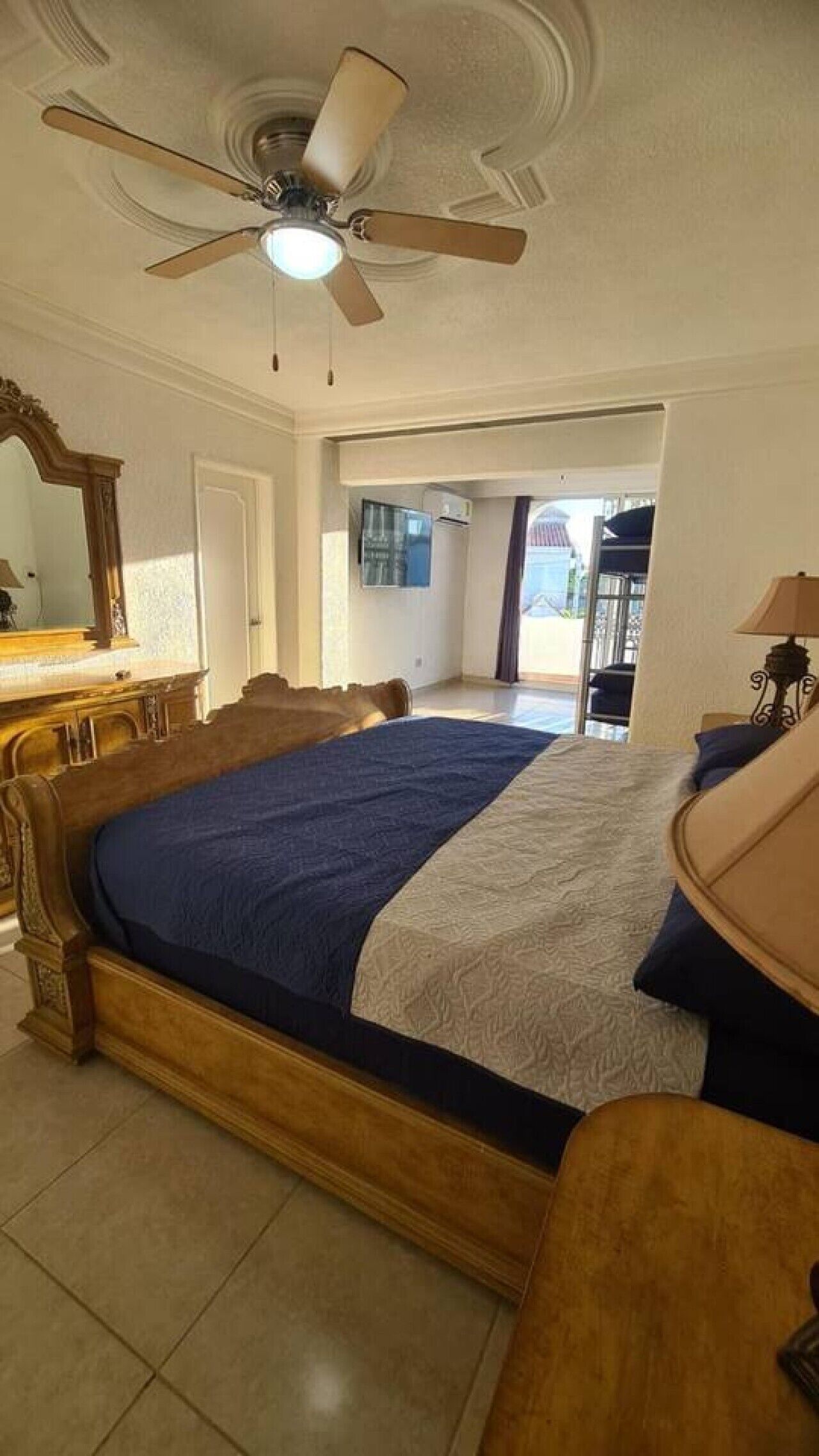 A bedroom with a large bed, a ceiling fan, a dresser, and a view of the outdoors.