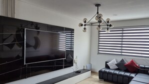 A modern living room with a large flat-screen TV mounted on the wall, a black leather sofa with cushions, and a chandelier. - Depa moderno piso7- Recamaras -Elevador+2 cajones (Zapopan)