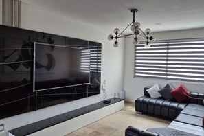 Ein modernes Wohnzimmer mit einem großen Flachbildfernseher an der Wand, einer schwarzen Ledersofa mit Kissen und einer Deckenleuchte.