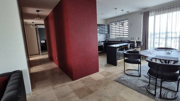 A modern kitchen with a central island, a dining area with a round table and chairs, and a red accent wall. - Depa moderno piso7- Recamaras -Elevador+2 cajones (Zapopan)