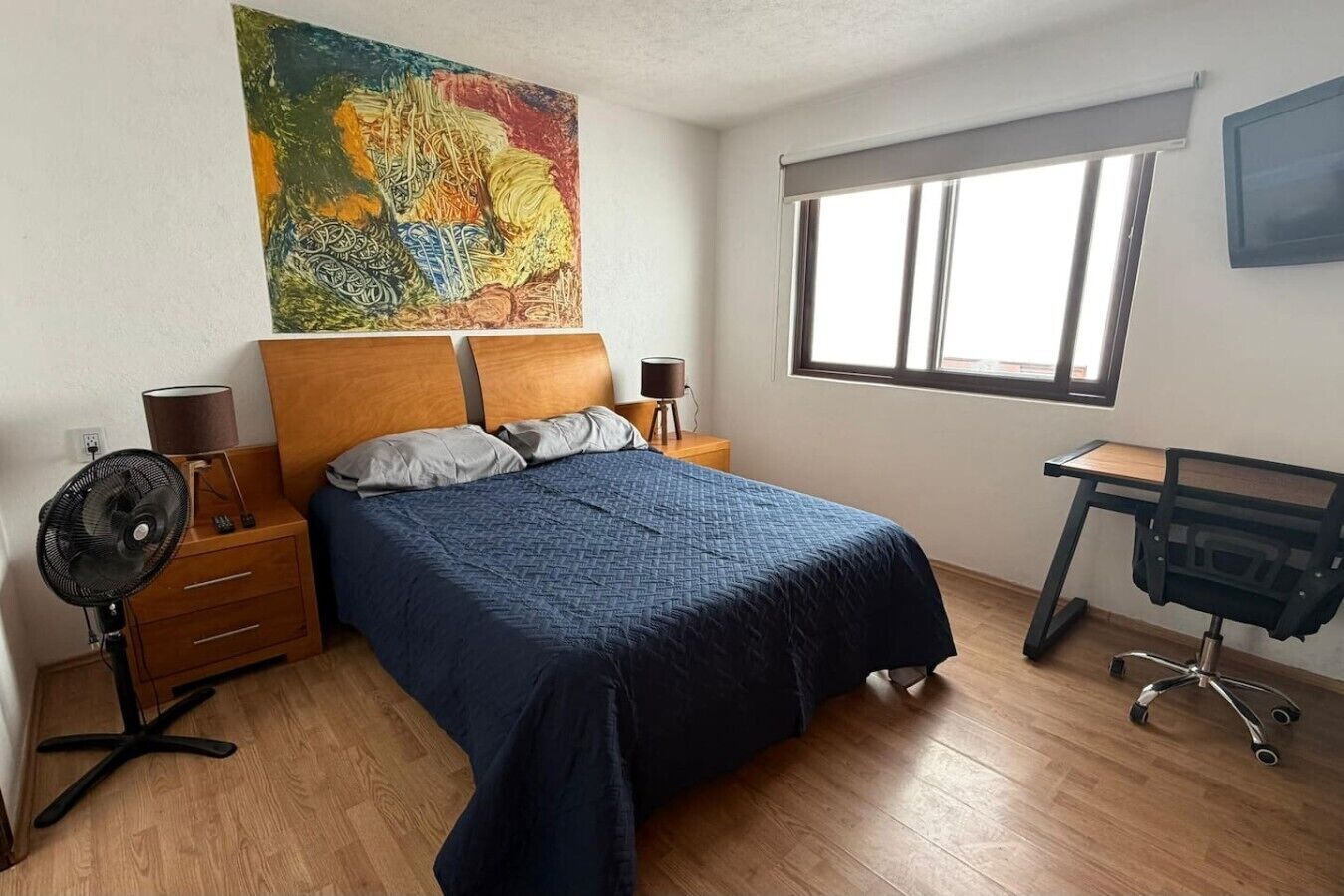 A bedroom with a bed, bedside lamps, a desk with a chair, a standing fan, and a large abstract painting on the wall.