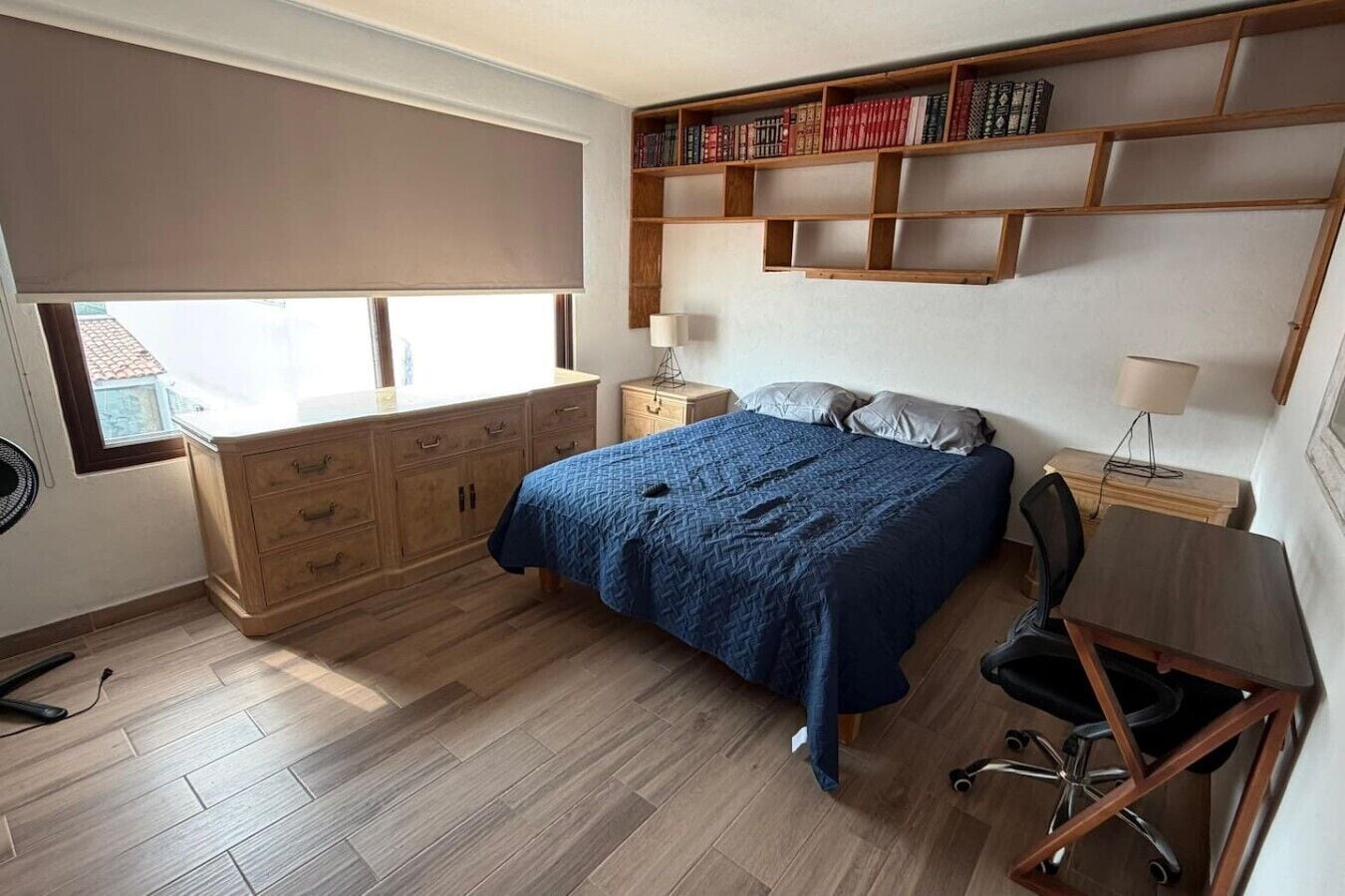 A bedroom with a bed, a desk, a chair, a bookshelf, and a window with a roller blind.
