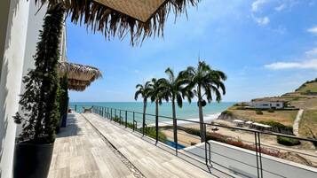 A balcony with a view of the beach, palm trees, and a thatched roof structure.