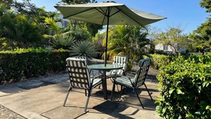 Een terras buiten met een tafel en stoelen, omringd door weelderige begroeiing en een grote parasol.