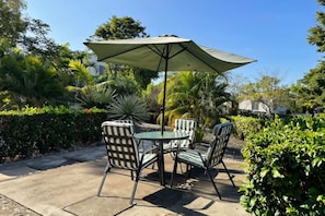 Un patio exterior con mesa y sillas, rodeado de abundante vegetación y un paraguas grande.