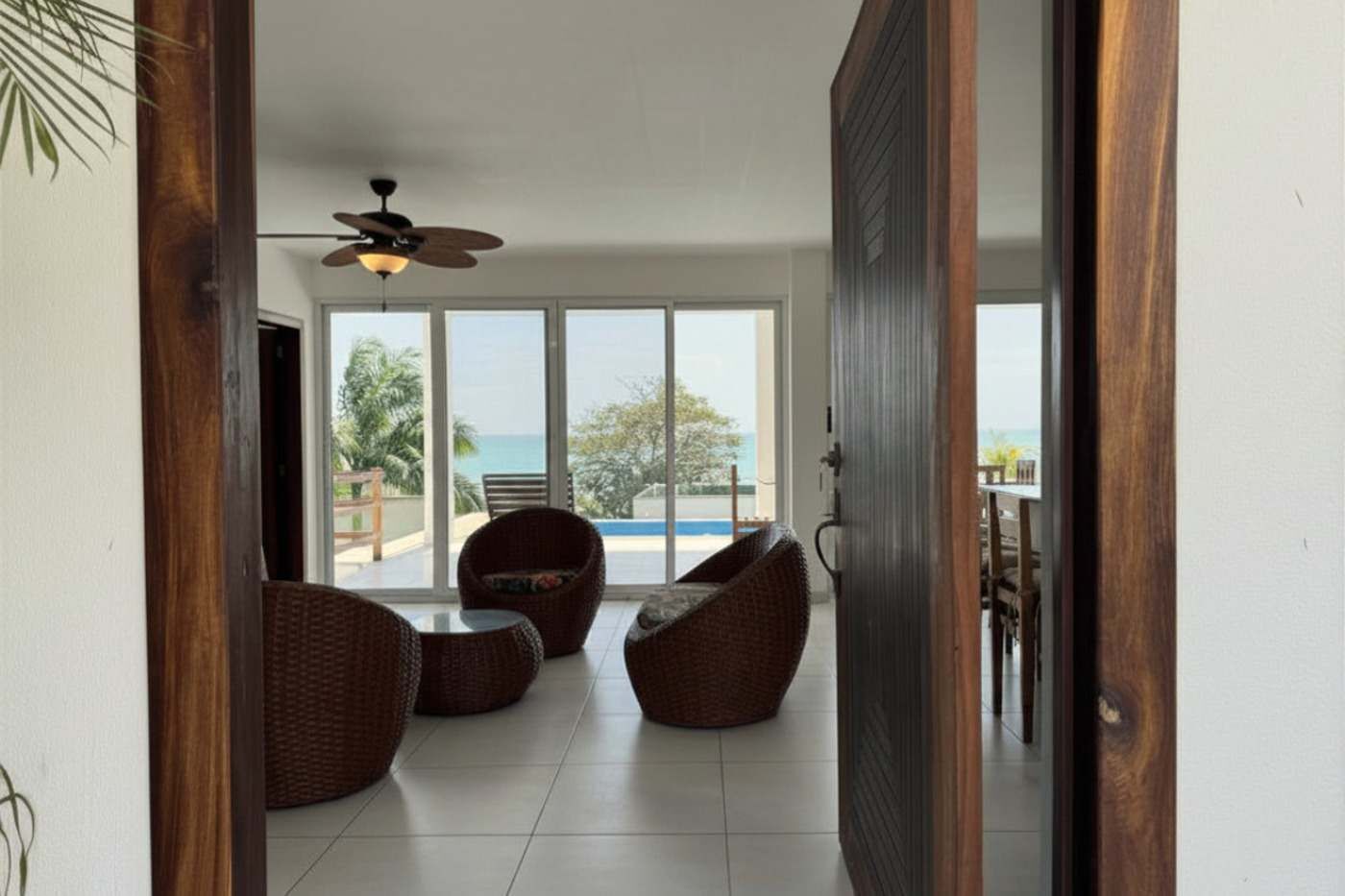 Una porta aperta su una stanza con ventilatore a soffitto, mobili in vimini e vista sul mare.