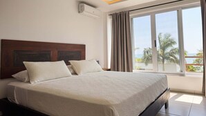 Ein Hotelzimmer mit einem großen Bett, einem Kopfteil, einem Fenster mit Vorhängen und Blick auf Palmen.