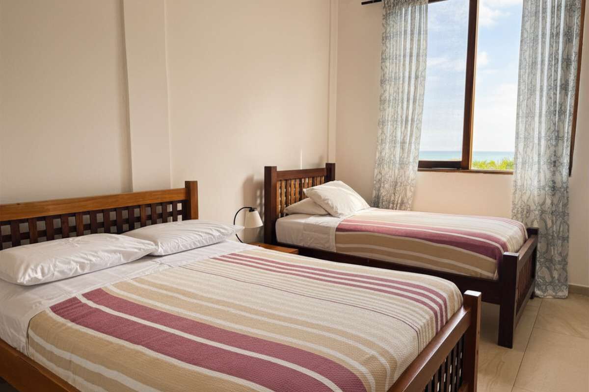 Quarto com duas camas de solteiro, janela com cortinas e vista para o mar.