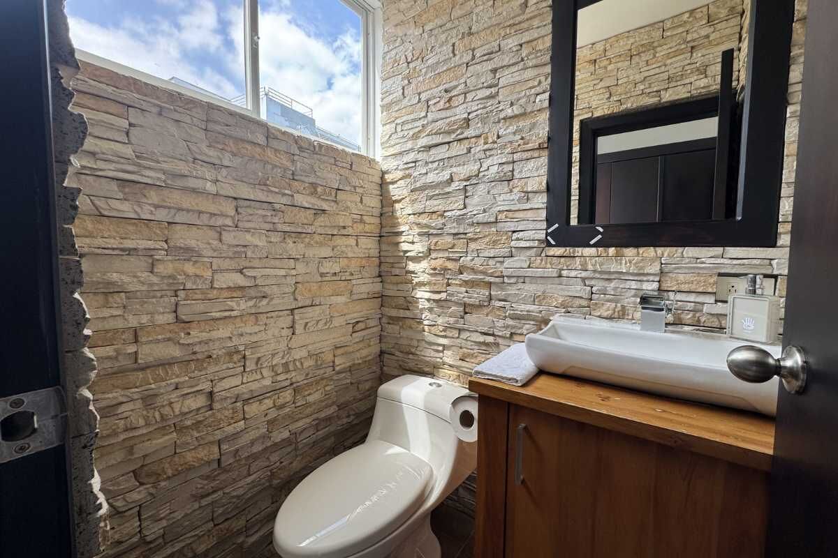 Un baño con pared de piedra, inodoro blanco, tocador de madera y espejo.