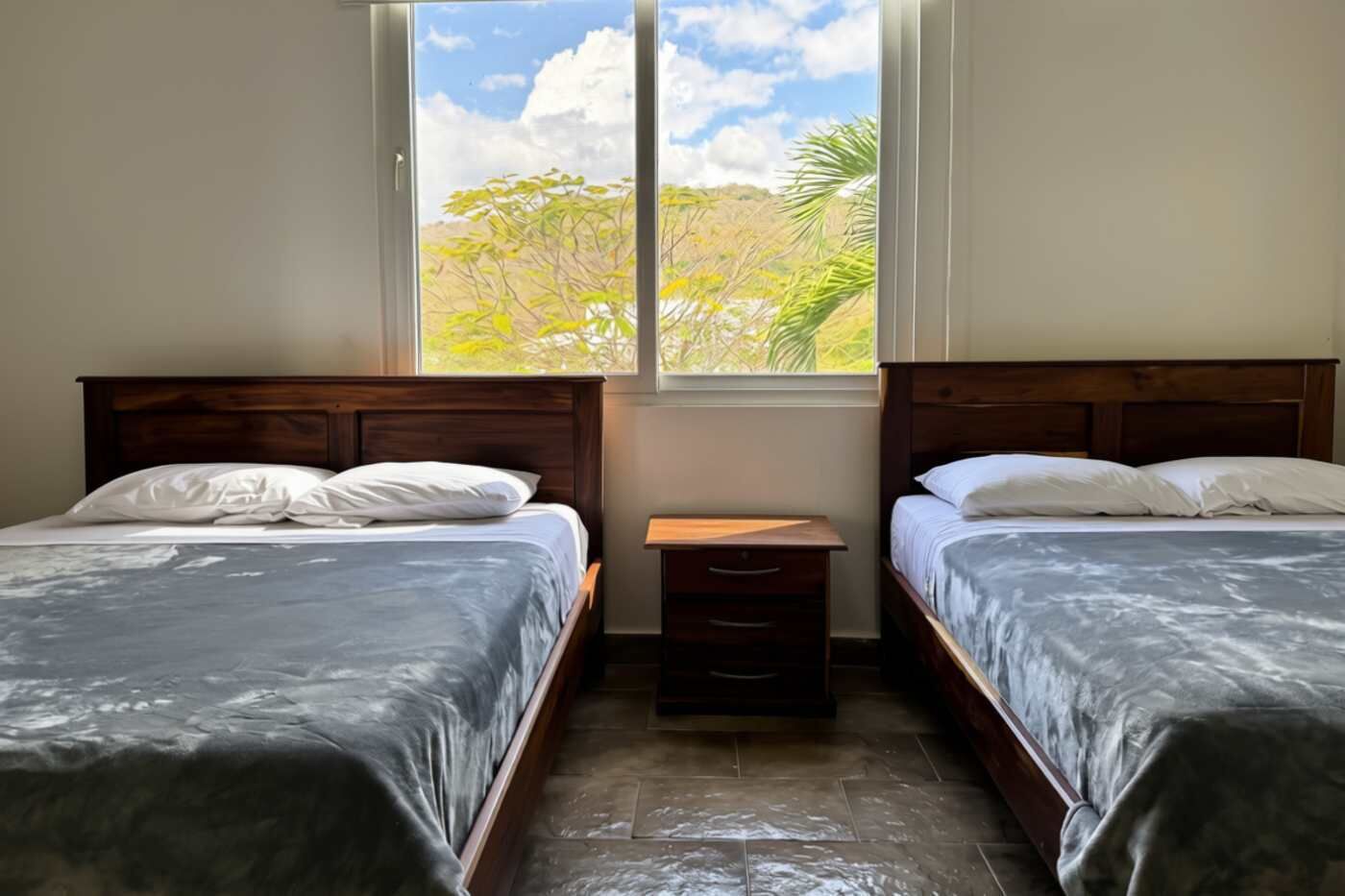 Una habitación con dos camas, una mesita de noche de madera y una ventana con vista a árboles.
