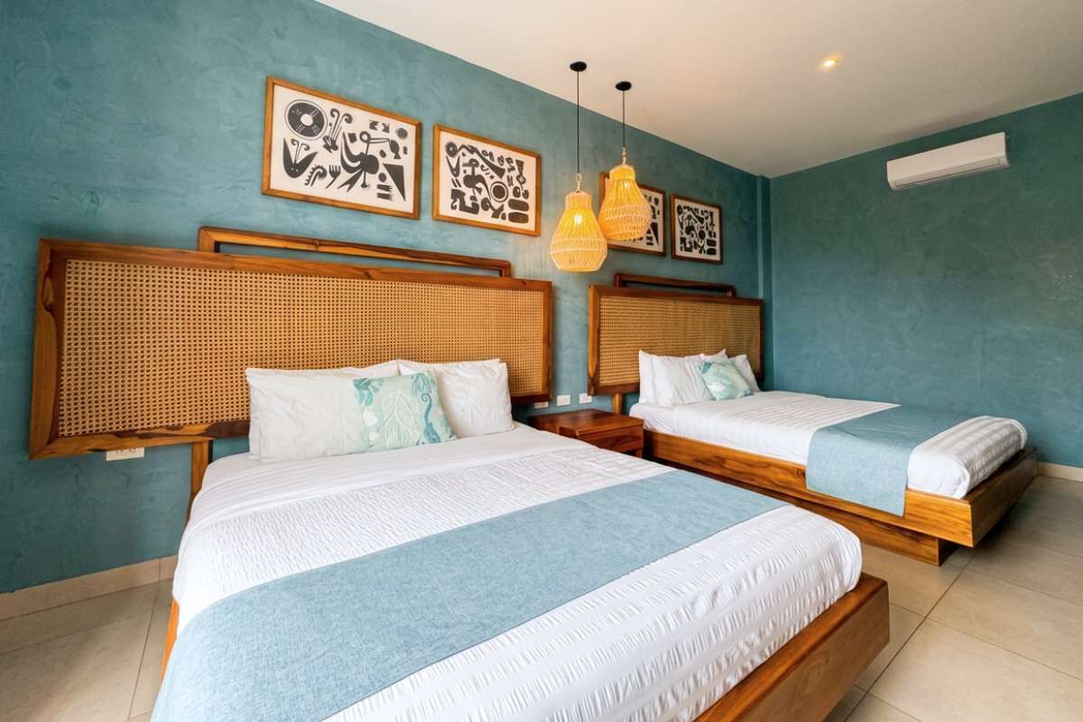 Habitación con dos camas, cada una con cabecera de madera y ropa de cama blanca. Se observan unidades de aire acondicionado fijadas a la pared y obras de arte enmarcadas sobre las camas.