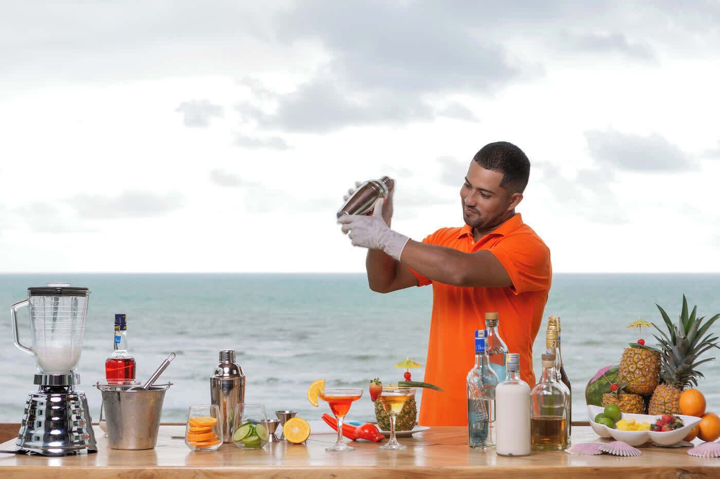 Una persona con una camisa naranja prepara bebidas en un bar con vista al océano.