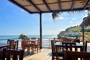 A covered outdoor seating area with wooden tables and chairs, overlooking a beach with a clear view of the ocean and a white building on the hillside.
