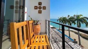 A balcony with a wooden bench, a large potted plant, and a view of the beach with palm trees and the ocean.