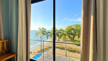 Una habitación con un amplio ventanal que da a una playa con palmeras y una piscina.