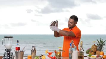Una persona con una camisa naranja prepara bebidas en un bar con vista al océano.