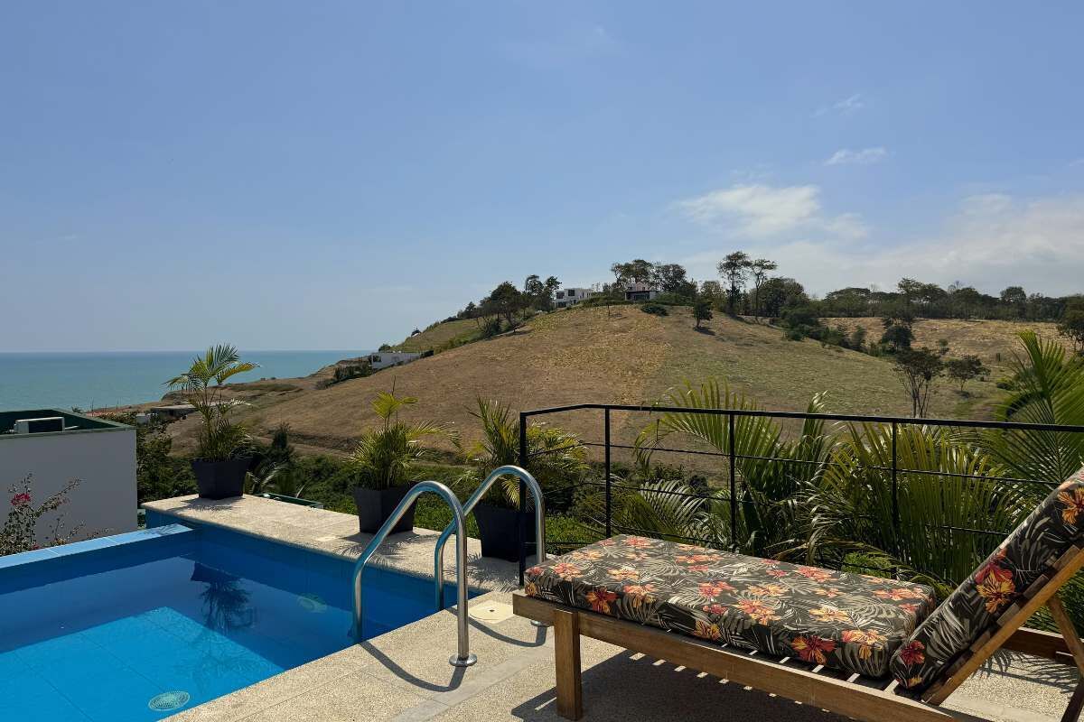 A pool area with a view of the ocean and a hill with trees.
