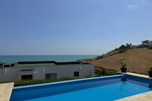 Una piscina moderna amb una vista clara de l'oceà i un paisatge amb turons al fons.