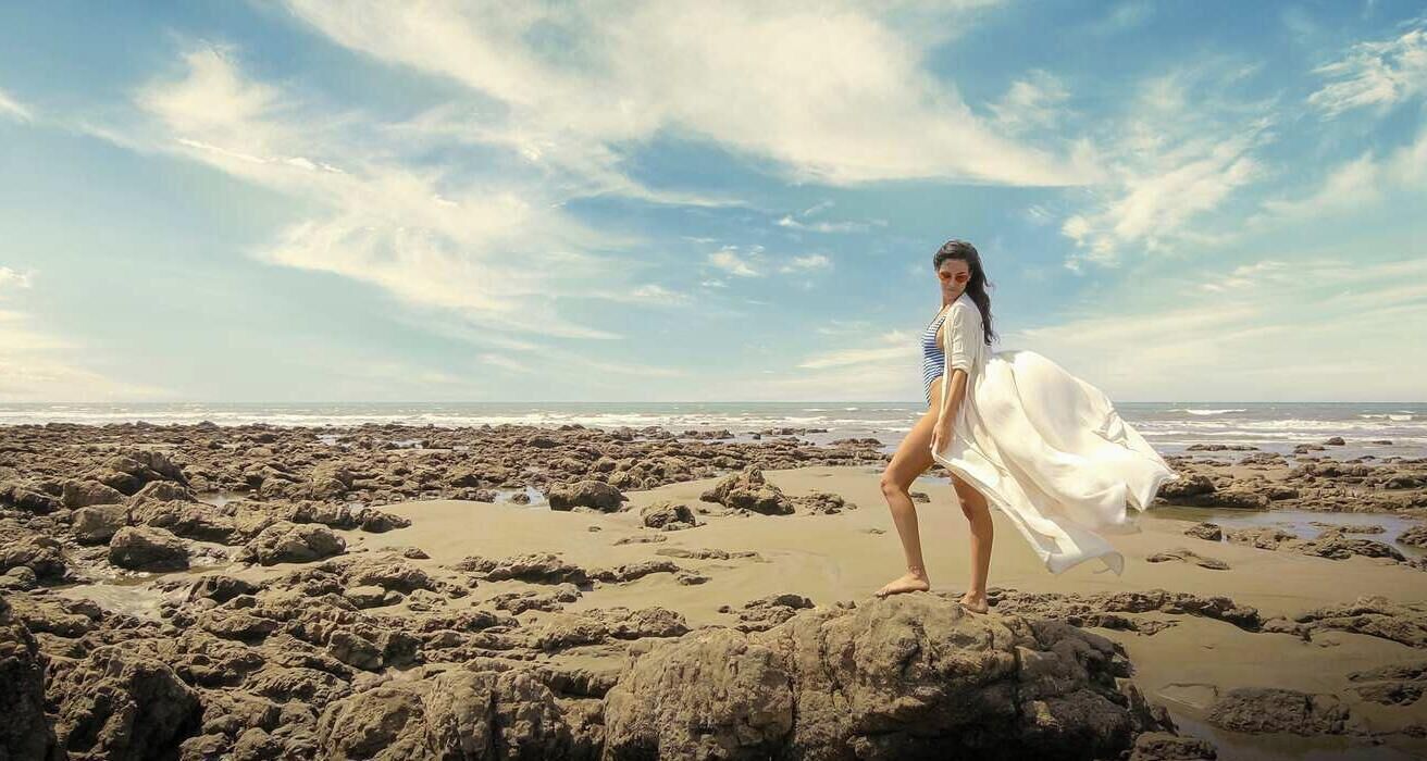 Una persona parada sobre terreno rocoso, con una playa y el océano al fondo.