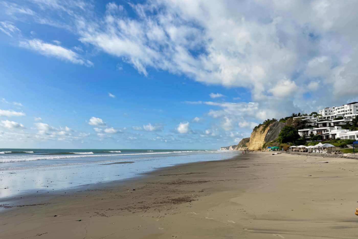 A beach with a clear sky, waves, and buildings on a cliff.