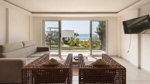 Ein modernes Wohnzimmer mit einer Couch, Korbstühlen, einem Flachbildfernseher und einem Balkon mit Meerblick.