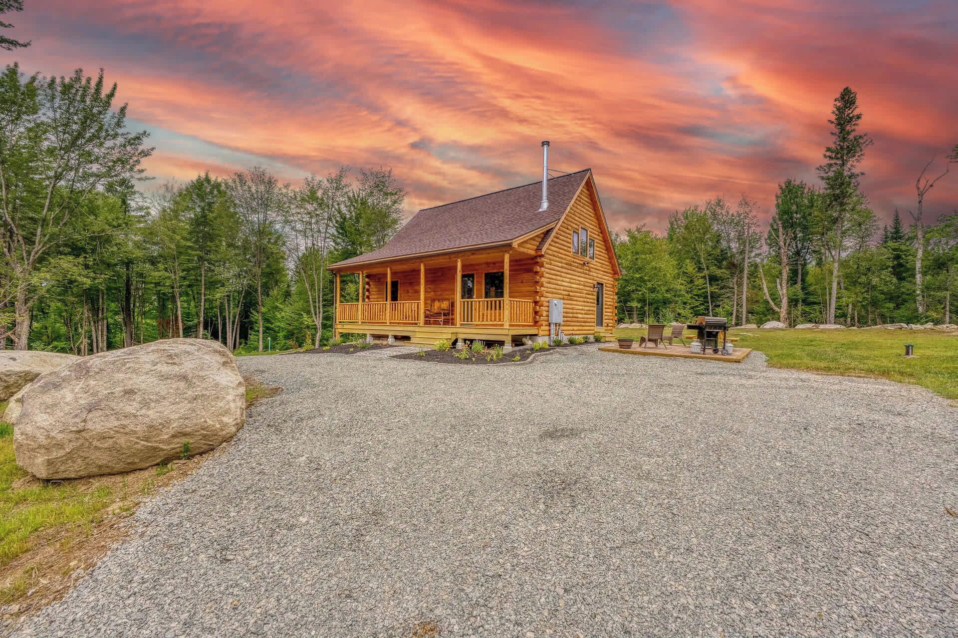 A log cabin with a porch and a chimney, surrounded by a gravel driveway and a forested area.