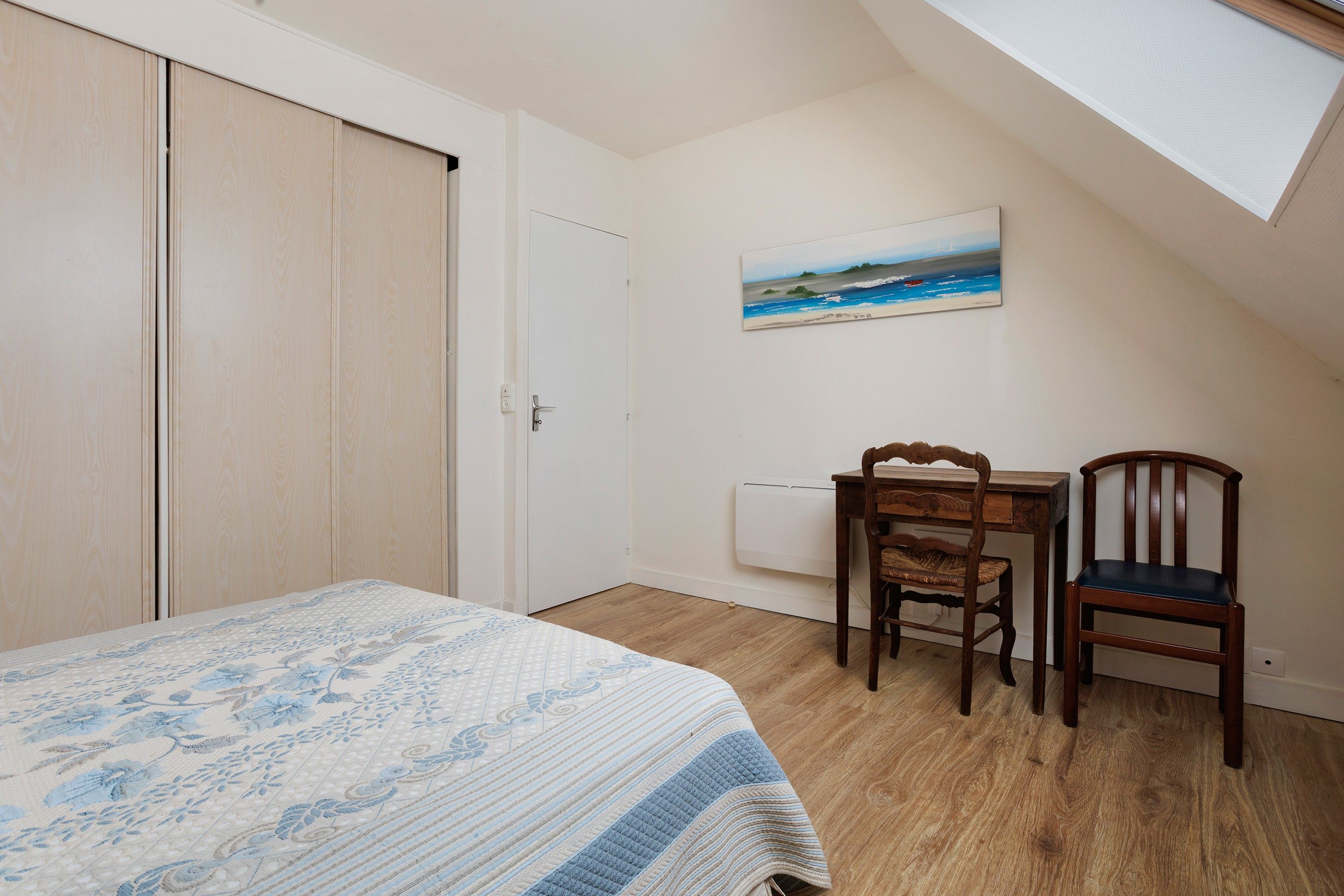 A bedroom with a bed, a desk, two chairs, a painting on the wall, and a sloped ceiling.