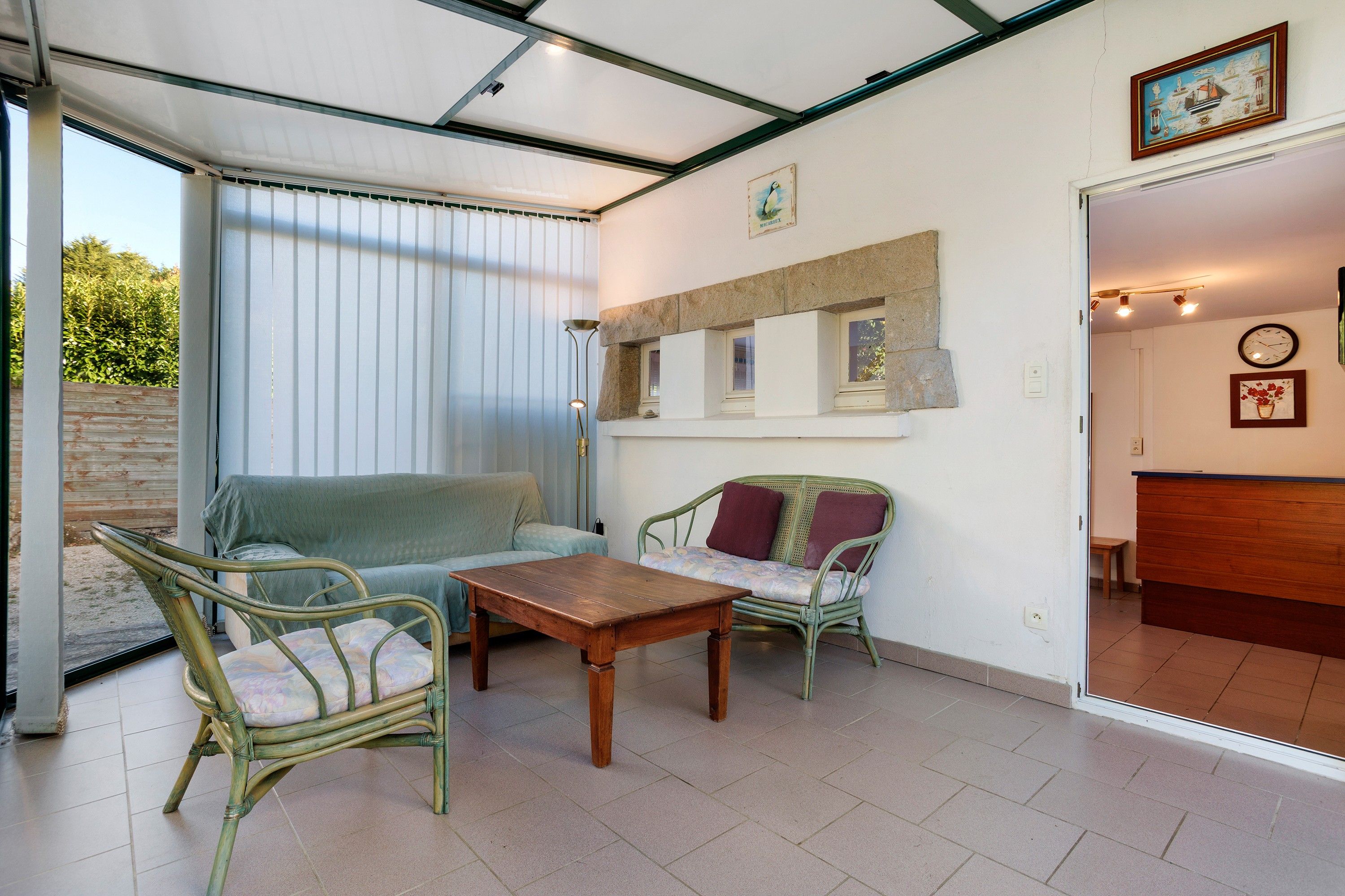 A room with a glass door leading to an outdoor area, a wooden table, two wicker chairs, a sofa, and a small wooden cabinet with decorative items.