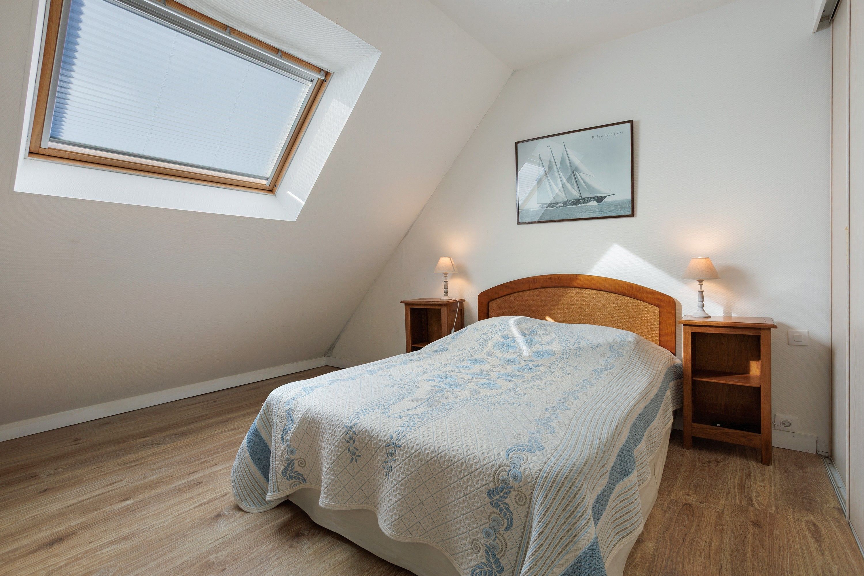 A bedroom with a sloped ceiling, a bed with a floral bedspread, two bedside tables with lamps, a framed picture on the wall, and a window with blinds.