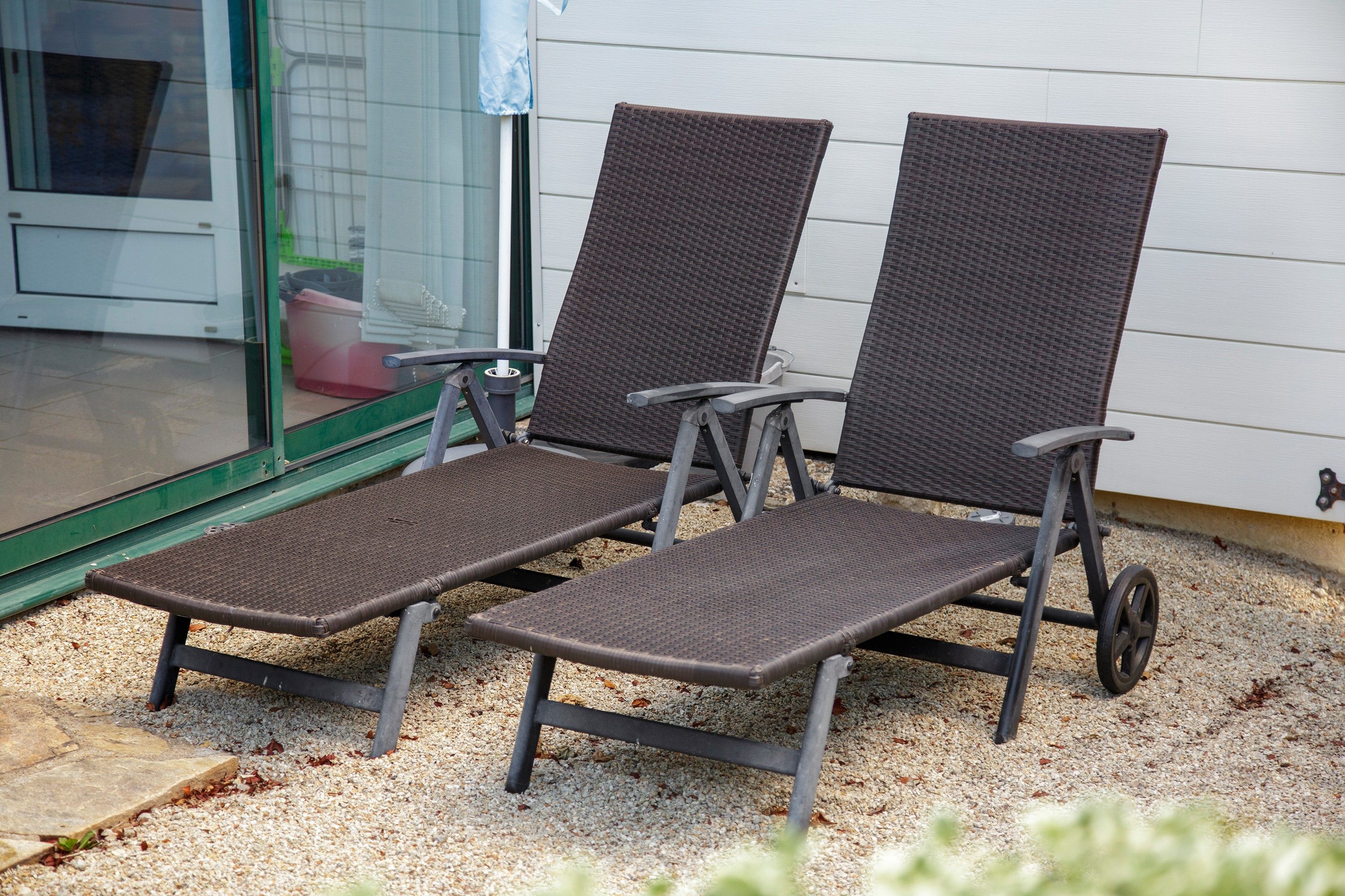 Twee loungesteunen met metalen frames en gevlochten zittingen, geplaatst op een grindpad voor een gebouw met een glazen deur.