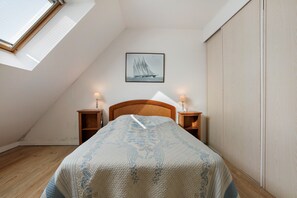 Une chambre avec un plafond en pente, un lit recouvert d’une couvre-lit à motifs floraux, deux tables de chevet avec des lampes, un tableau encadré représentant un voilier et une armoire.