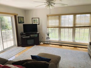 A living room with a flat-screen TV on a stand, a ceiling fan, and a large window with blinds. - 2 blocks to Roger Wheeler Beach 4-bedroom house perfect for your Summer getaway! (Narragansett)