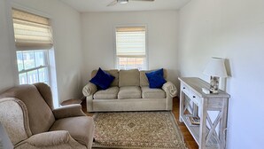 En stue med en beige sofa, en blå lænestol, trægulv, loftventilator og et sofabord med lampe.