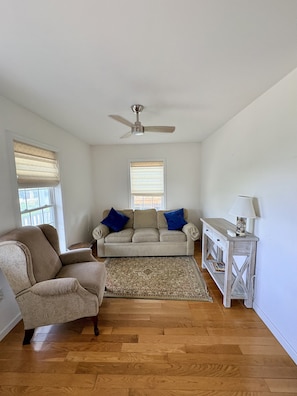 A living room with a beige sofa, a blue armchair, a wooden floor, a ceiling fan, and a side table with a lamp. - 2 blocks to Roger Wheeler Beach 4-bedroom house perfect for your Summer getaway! (Narragansett)