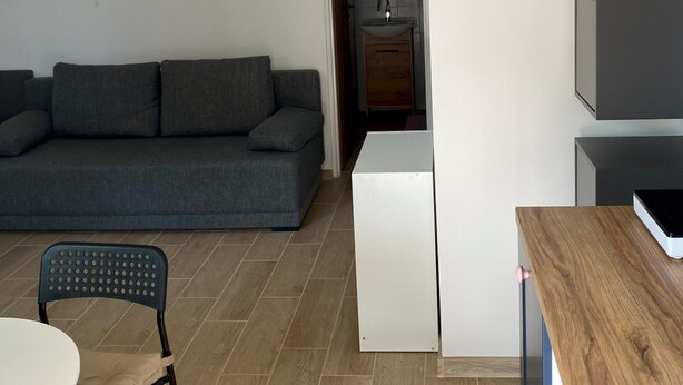Moderni olohuone, jossa on harmaa sohva, pieni ruokapöytä, tuoli sekä pieni, käytännöllinen keittiö, jossa on pesuallas ja kaappeja.