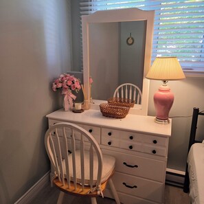 Une coiffeuse blanche avec un miroir, une lampe rose, un panier en osier et une chaise.