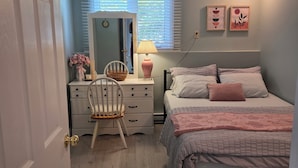 A bedroom with a bed, bedside table, lamp, chair, dresser, and a window with blinds. - Zen Cove- Home in West Saint John. 7 minutes from the Irving Nature Park. (Saint John)
